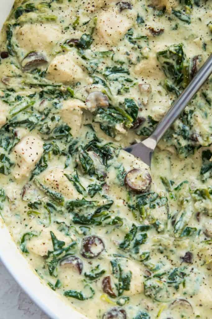 A pan of paleo chicken Alfredo with spinach and olives with a spoon in it