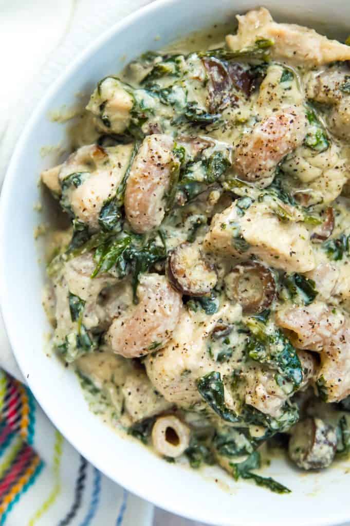 A bowl of paleo chicken Alfredo with spinach and olives over noodles.