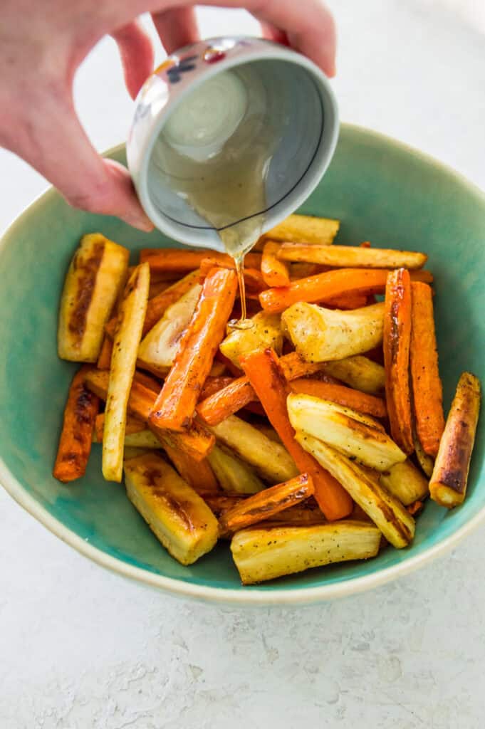 A honey glaze being poured over a bowl filled with roasted carrots and parsnips.