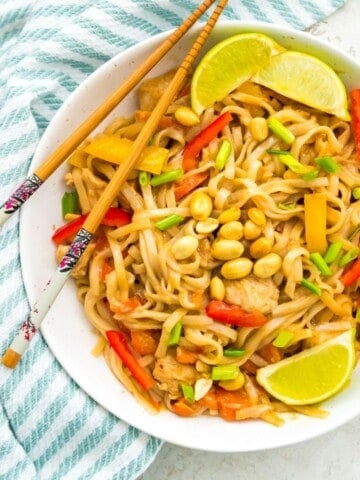 A bowl of chicken pad Thai topped with peanuts and garnished with lime wedges.