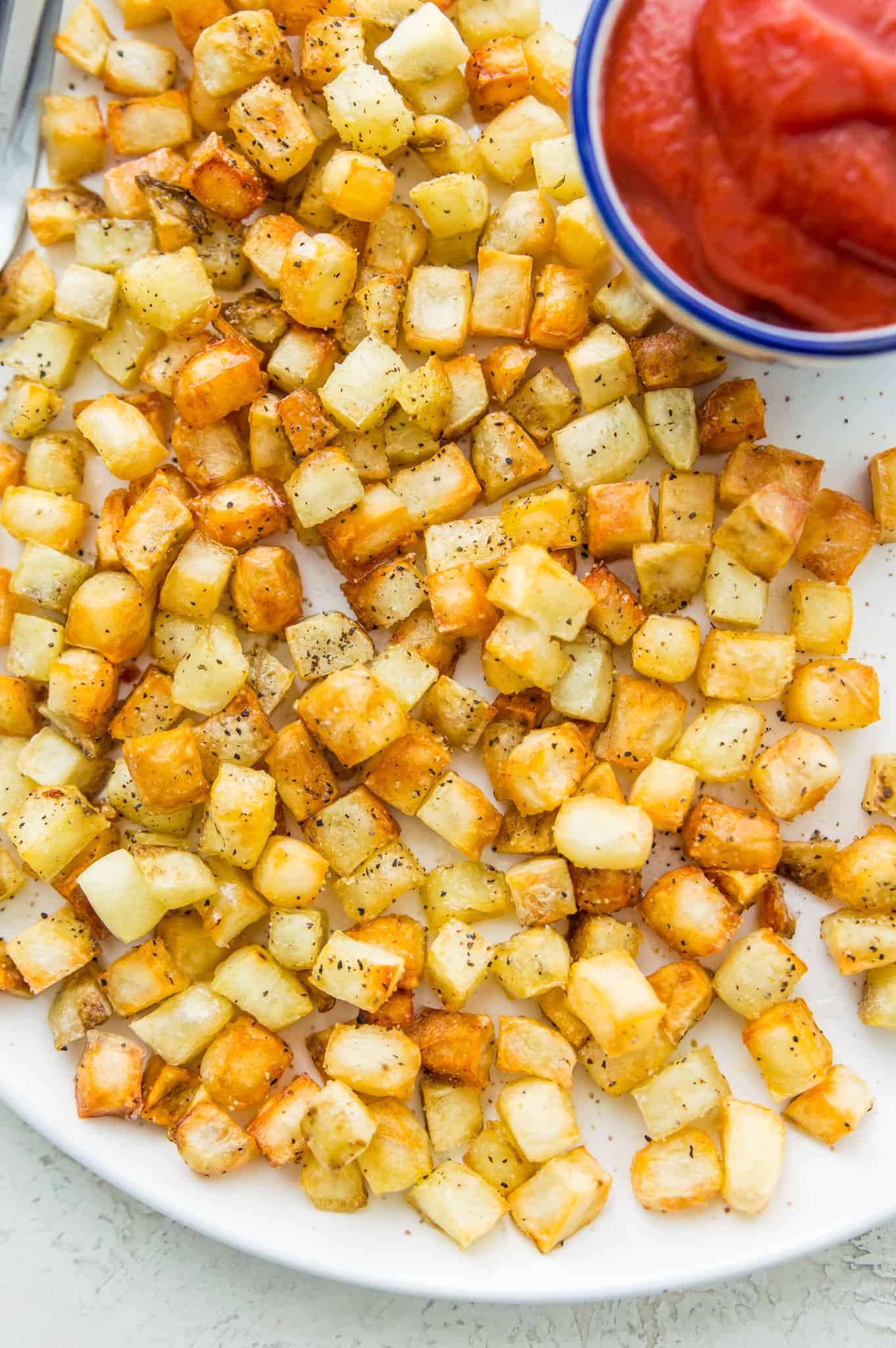 A plate of hash browns seasoned with black pepper with a side dish with ketchup in it.