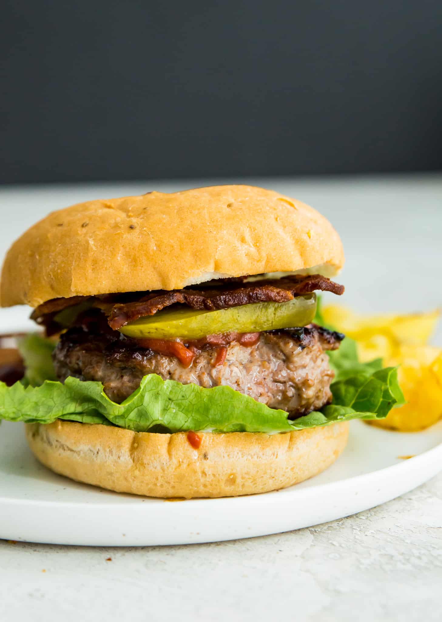 A duck burger on a plate in a bun with chips beside the burger and the burger is garnished with pickles, lettuce and bacon.