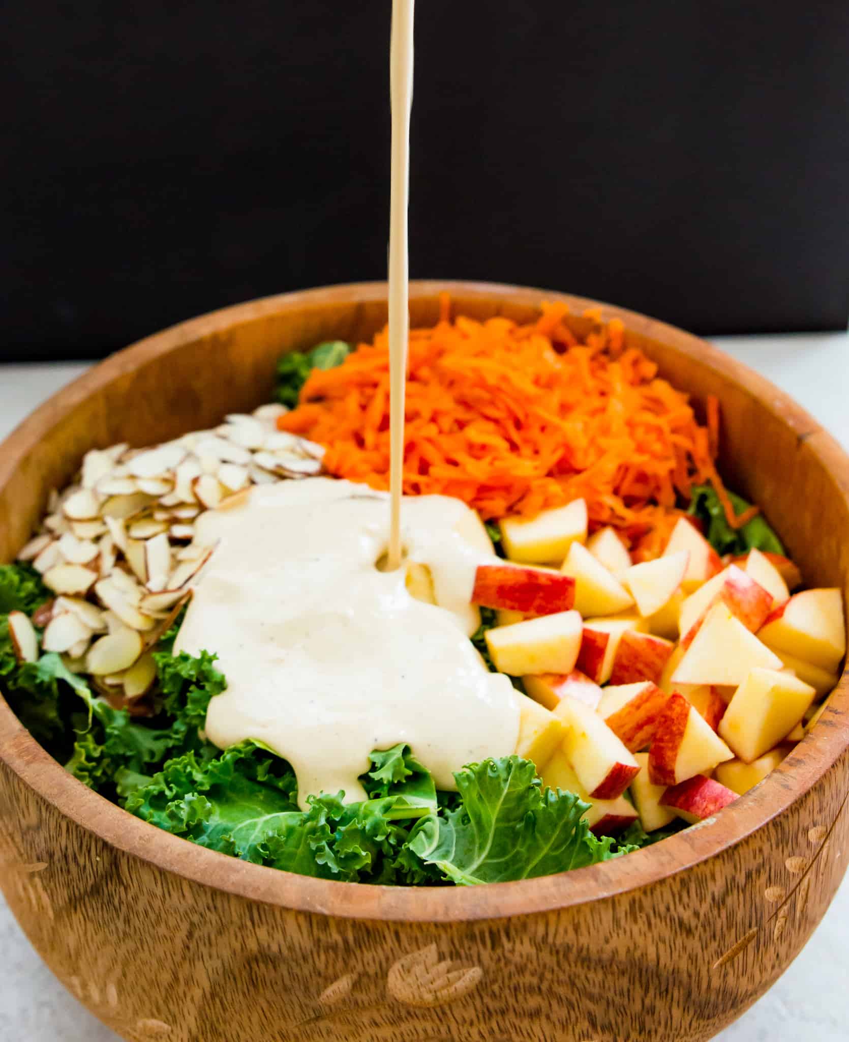 A creamy dressing being poured over a wooden mixing bowl full of kale, almonds, carrots, and apples.