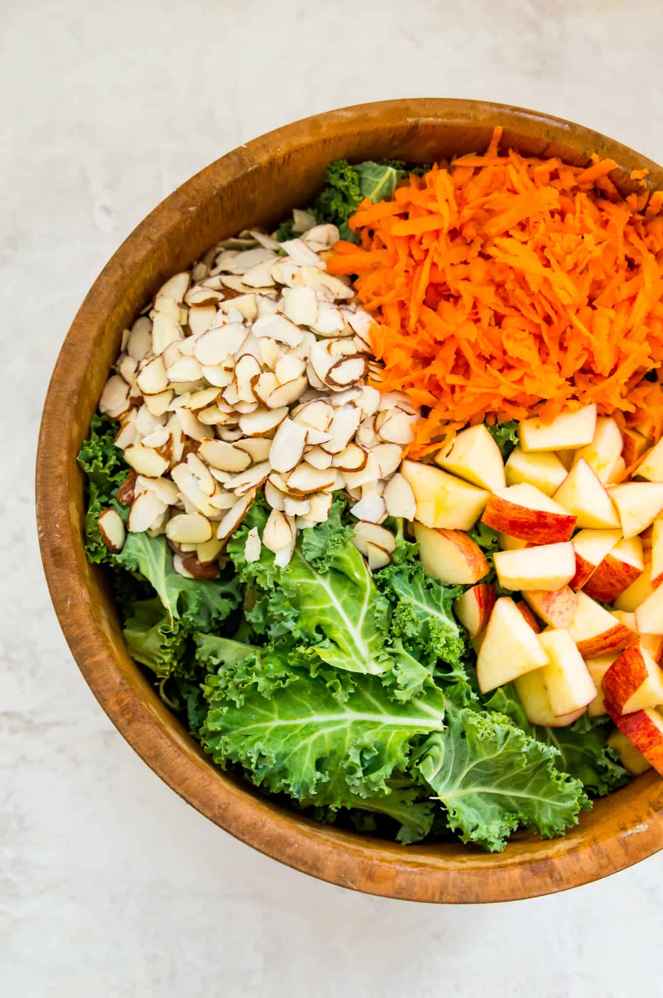 A wooden salad bowl filled with chopped kale, slivered almonds, chopped apples and shredded carrots.