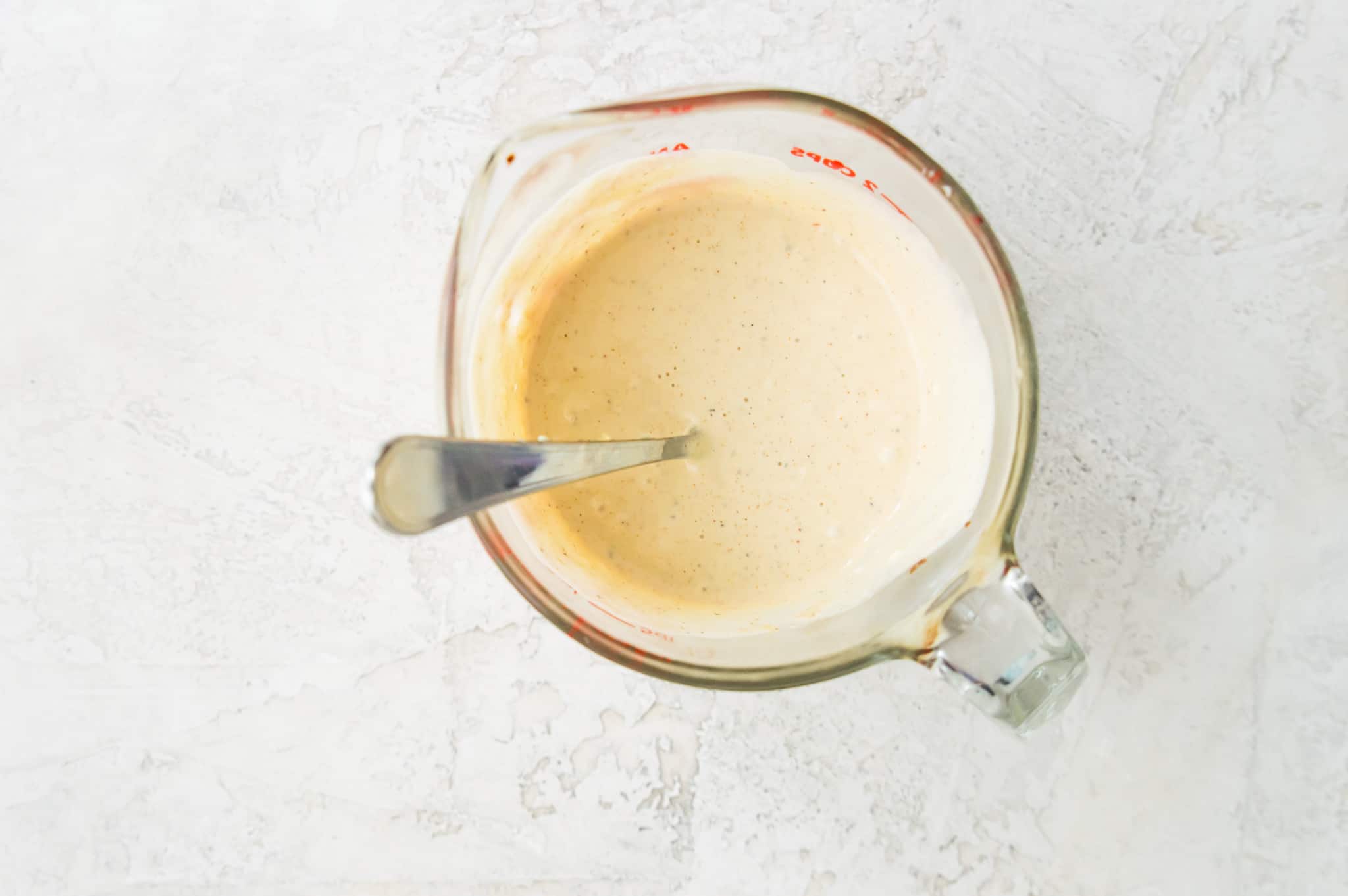 A glass measuring cup filled with a creamy salad dressing with a spoon in it.