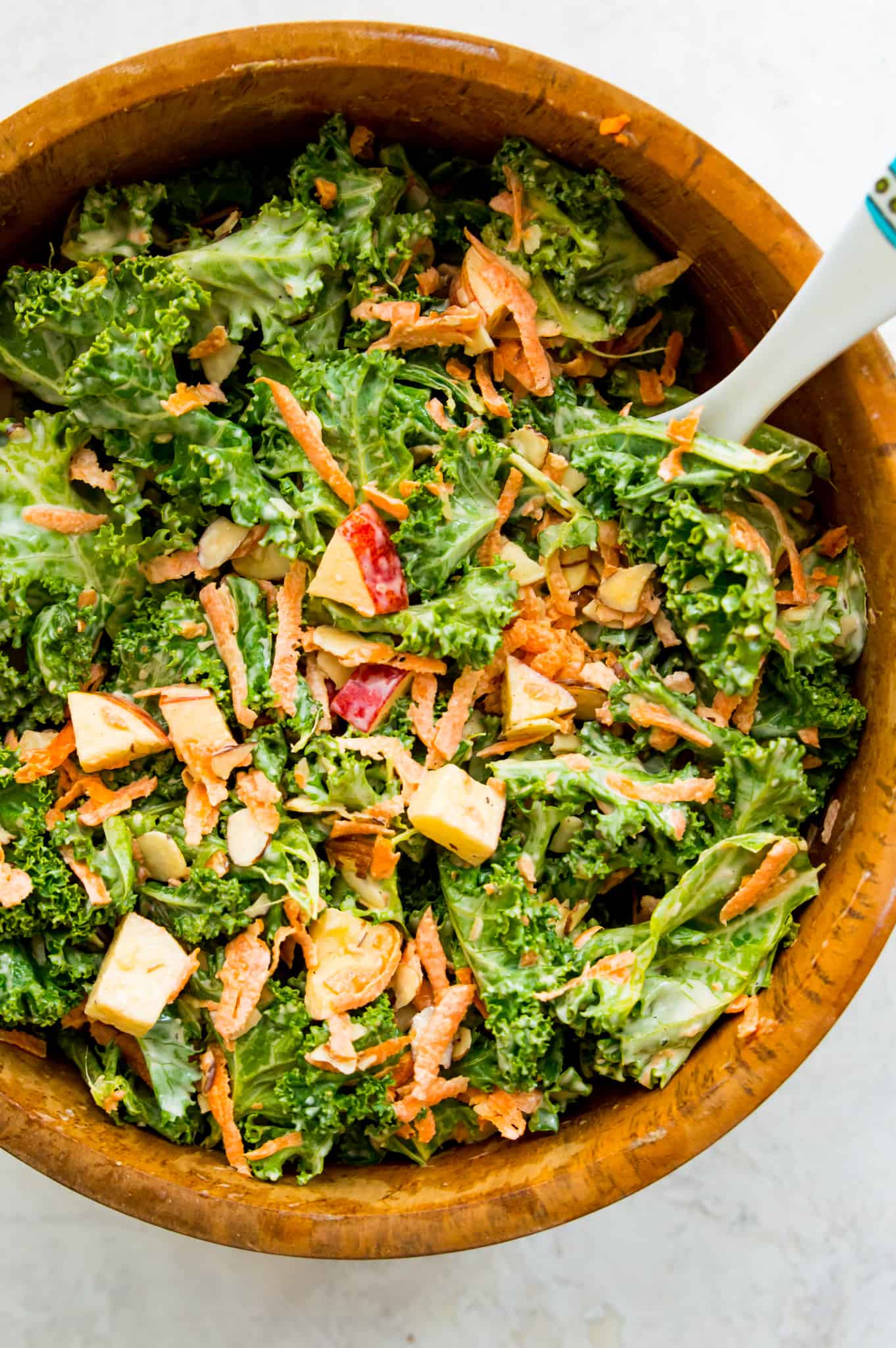 Pieces of red apple, carrots, slivered almonds, and fresh kale in a wooden mixing bowl with dressing.