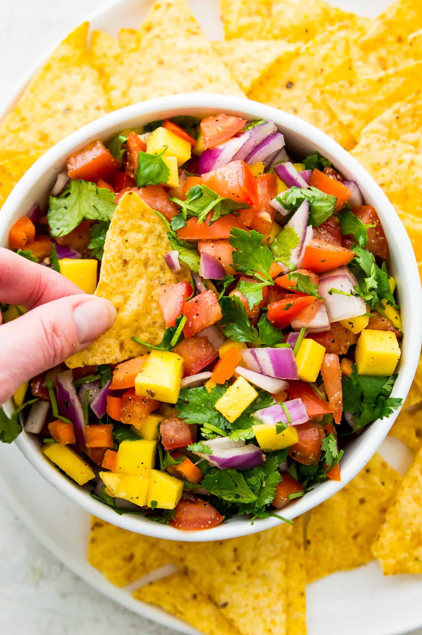 A hand dipping a corn tortilla chip into a bowl of mango salsa with tomatoes and red onions.