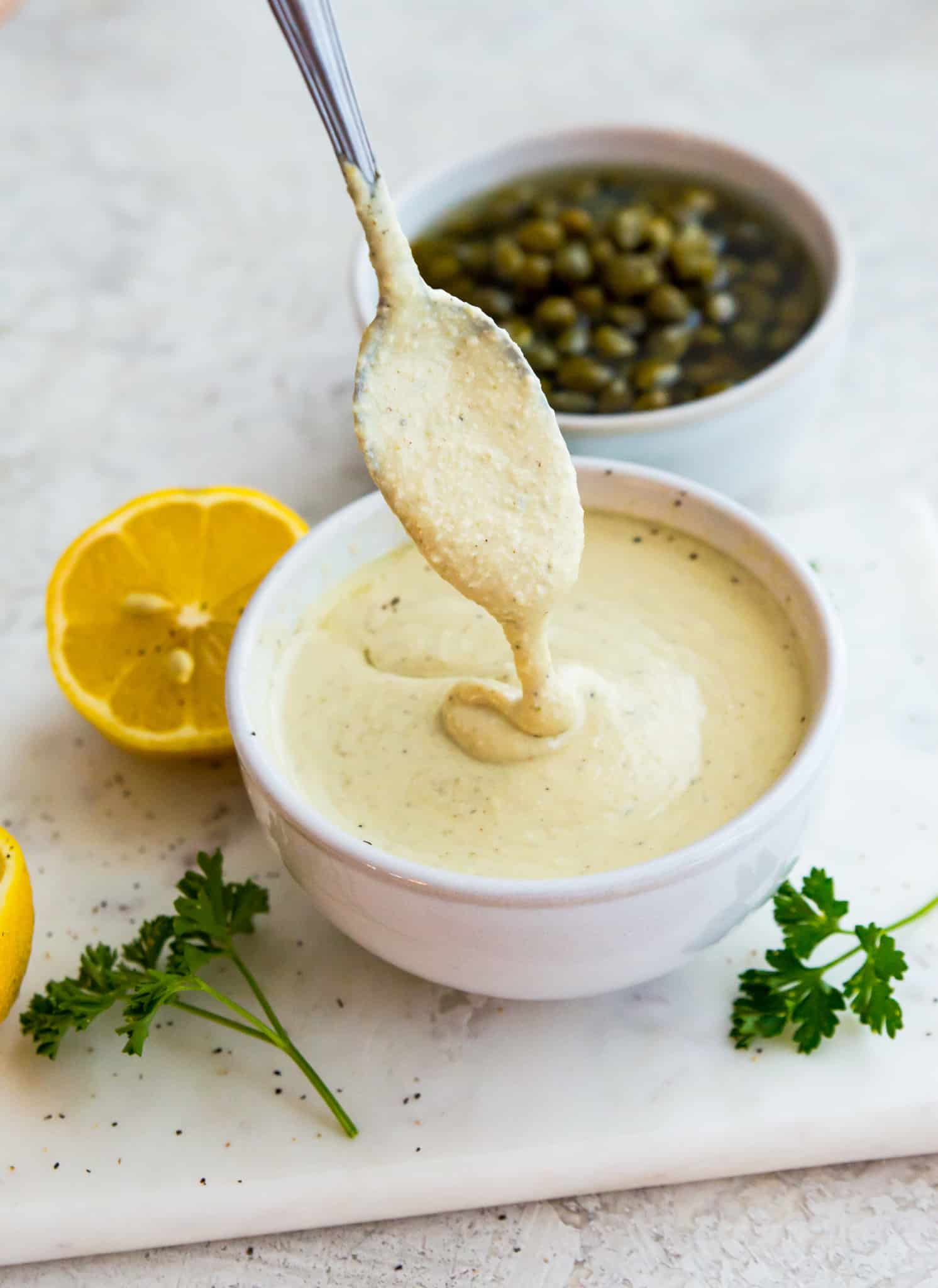 A bowl of vegan Caesar dressing with a spoon in it and surrounded by a lemon wedge and fresh parsley.