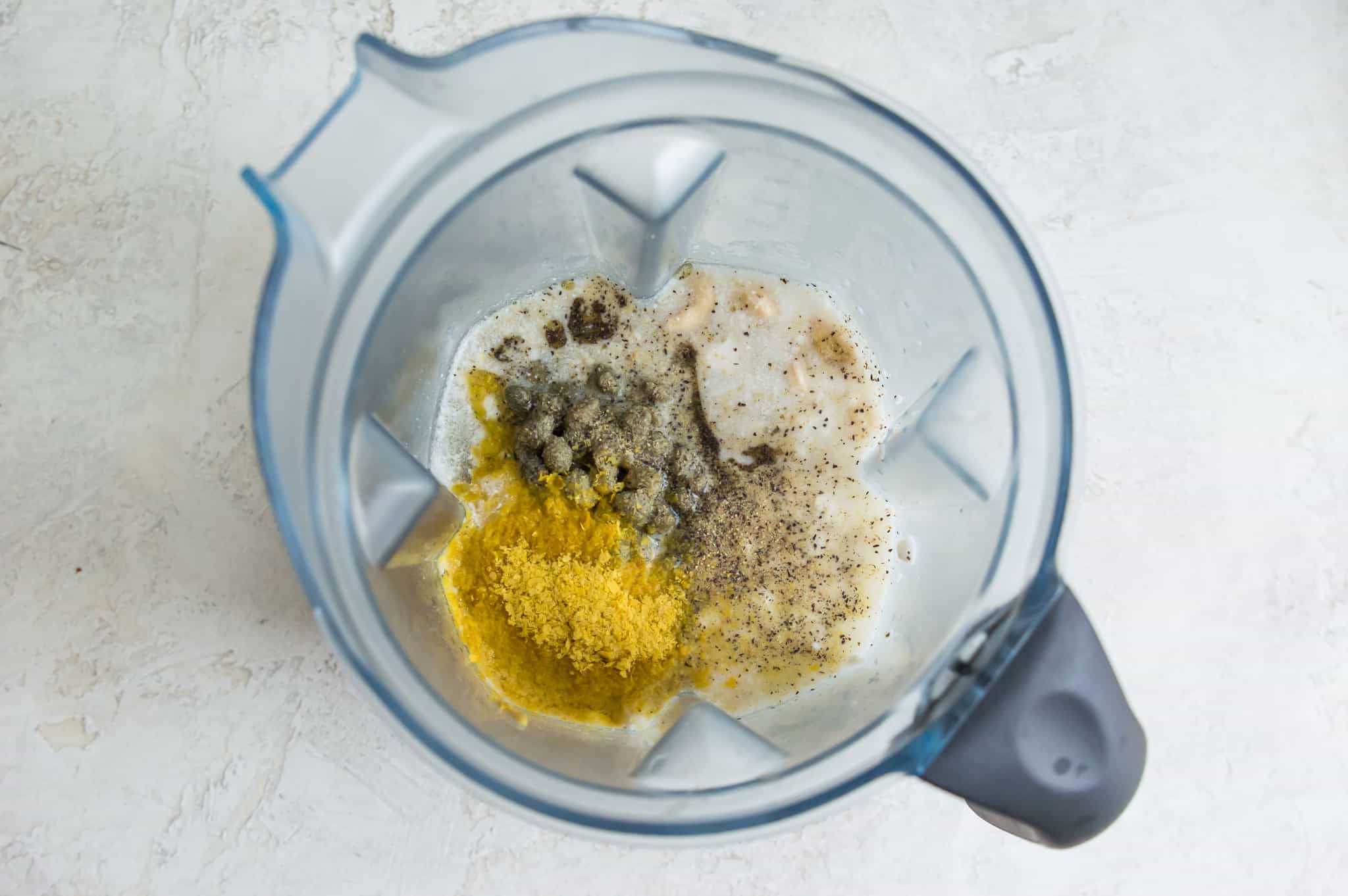 A blender filled with nutritional yeast, coconut milk, capers, water and spices. 