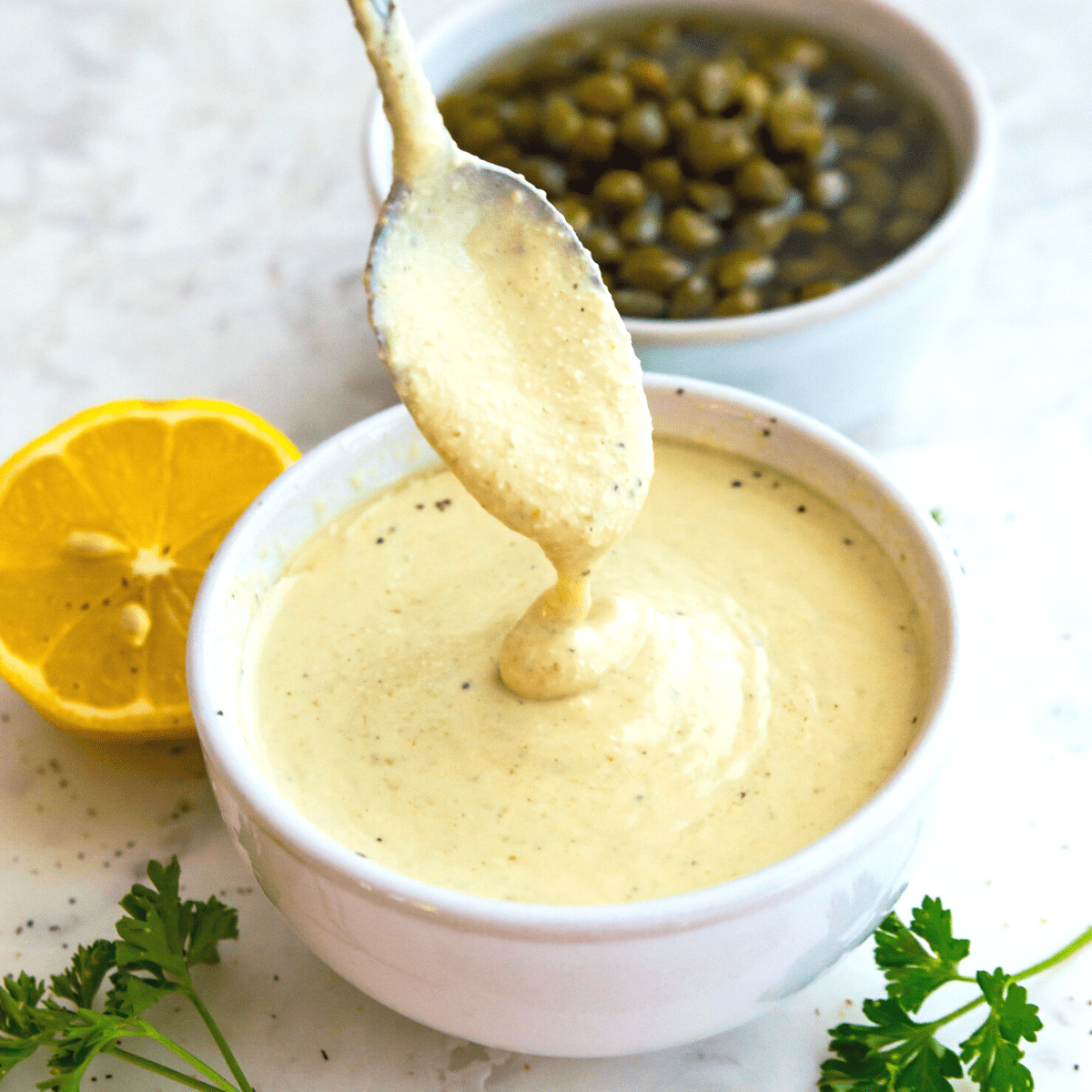 A bowl of vegan caesar dressing with a spoon in it and surrounded by a lemon wedge, fresh parsley and capers. 