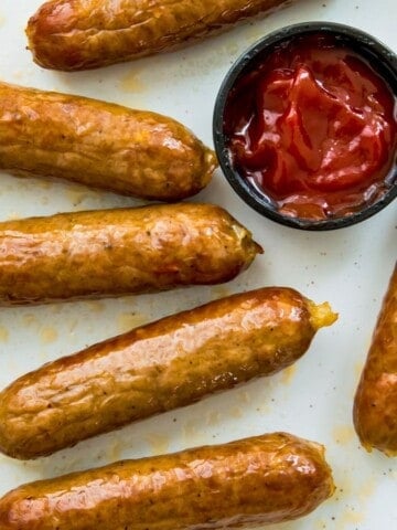 A plate of cooked bratwurst sausages with a dish of ketchup beside them.