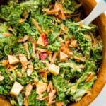 A wooden salad bowl filled with a kale apple salad with a serving spoon in it.