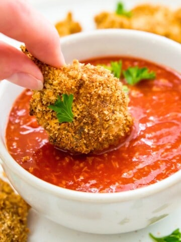 A breaded vegan shrimp being dipped into a bowl of shrimp cocktail sauce.