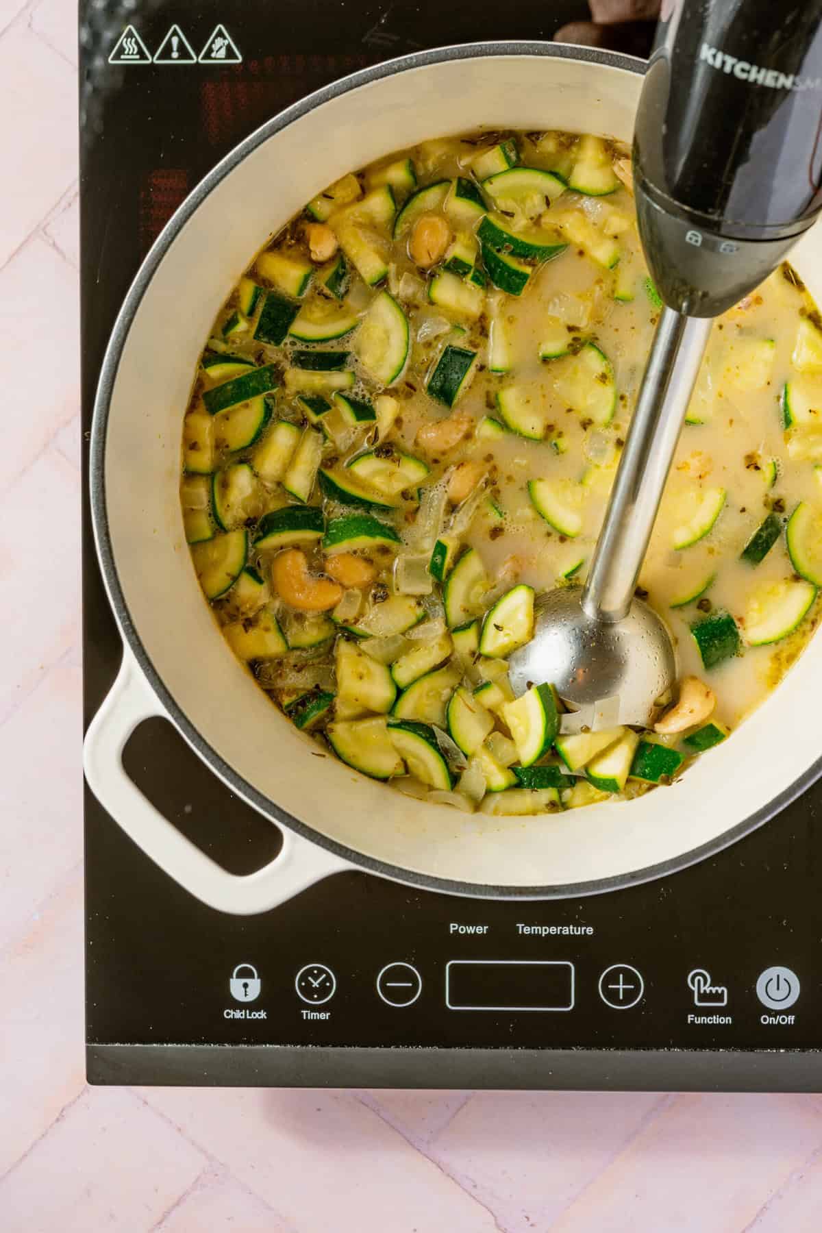 An immersion blender blending a pot full of coconut milk, chopped zucchini and cashews.