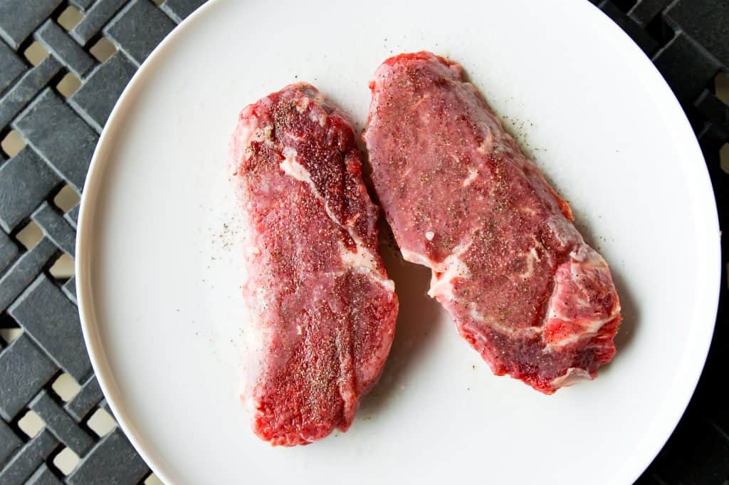 Two frozen steaks on a plate coated in salt and pepper.