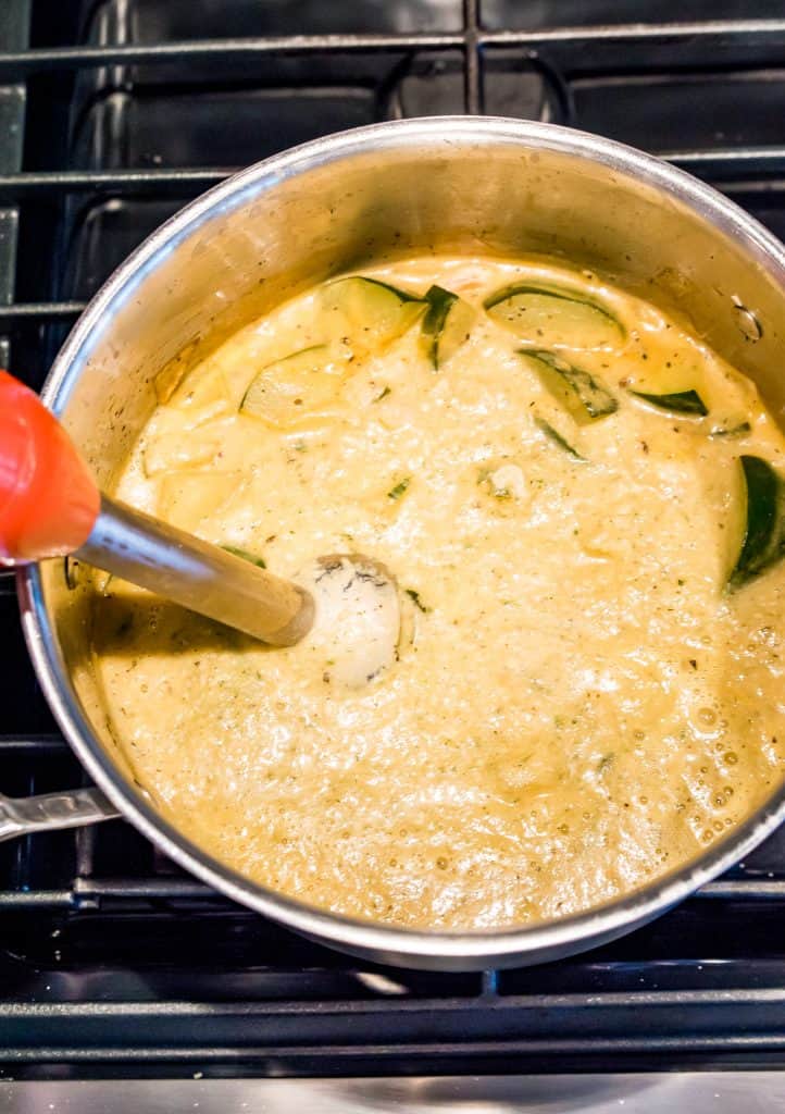 A large pot on the stovetop filled with zucchini soup and an immersion blender in it.