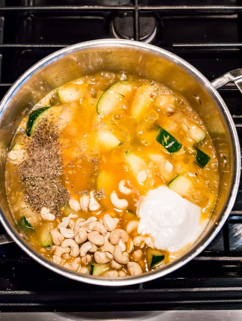 A large pot on the stove with zucchini, coconut milk, cashews and spices cooking in it.