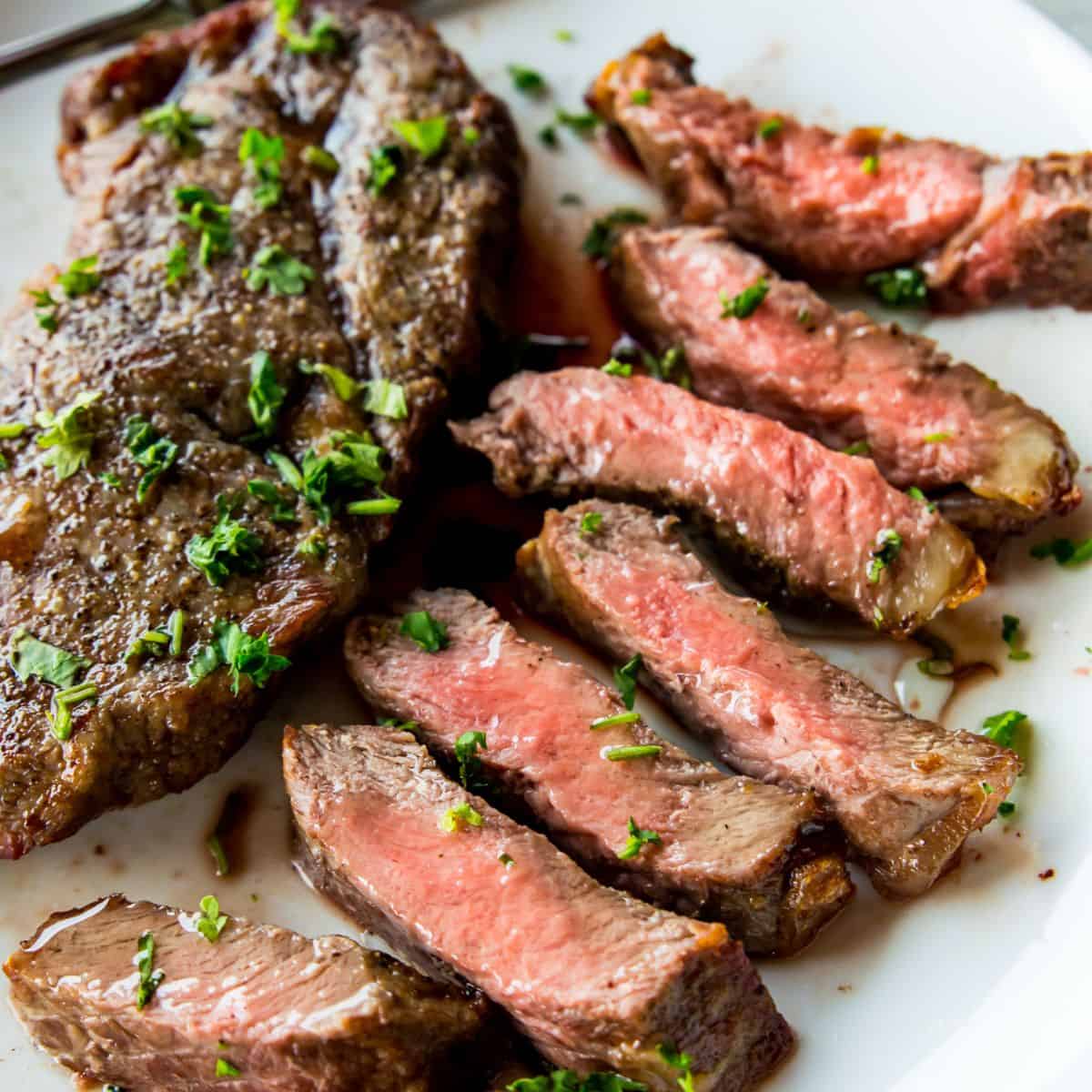 A piece of cut up steak on a plate with a full piece of steak beside it.