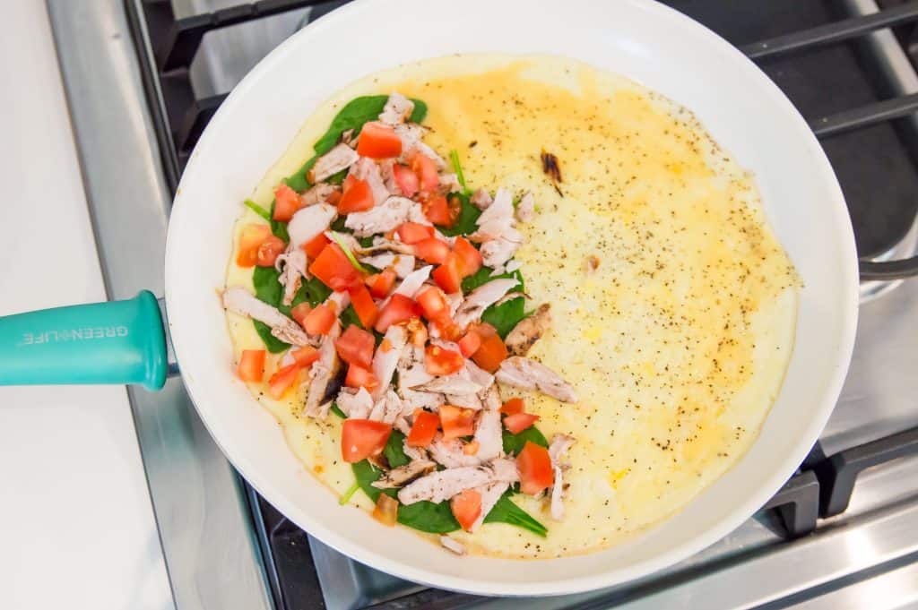 An omelette cooking in a pan filled with chopped chicken, spinach, and tomatoes. 