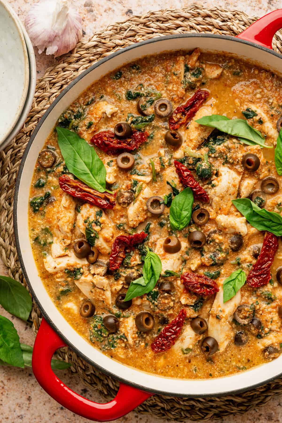 A pan with a sun-dried creamy tomato chicken dish in it topped with black olives and fresh basil.