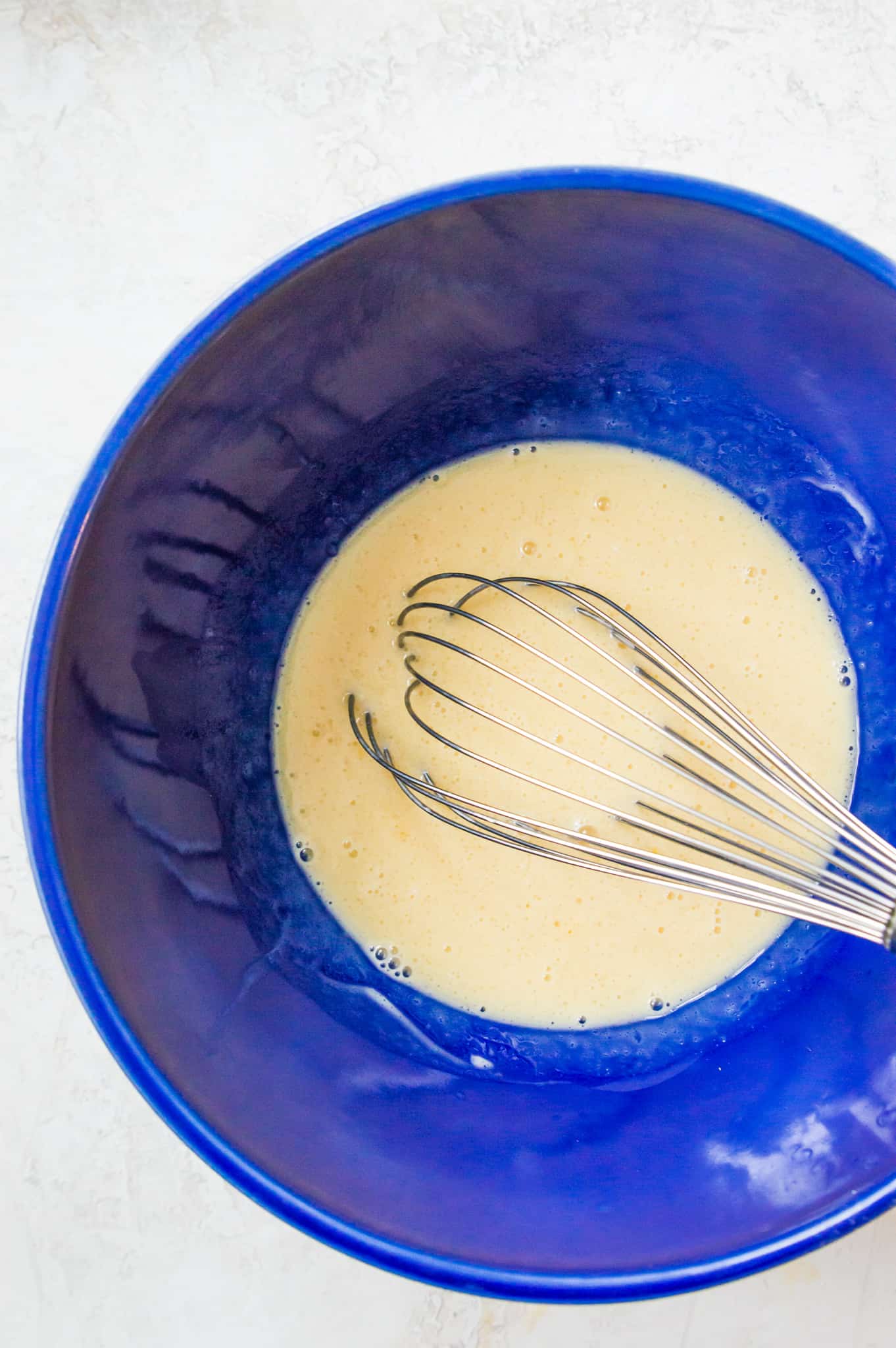 A large blue bowl filled with whisked egg and milk with the whisk in the bowl.