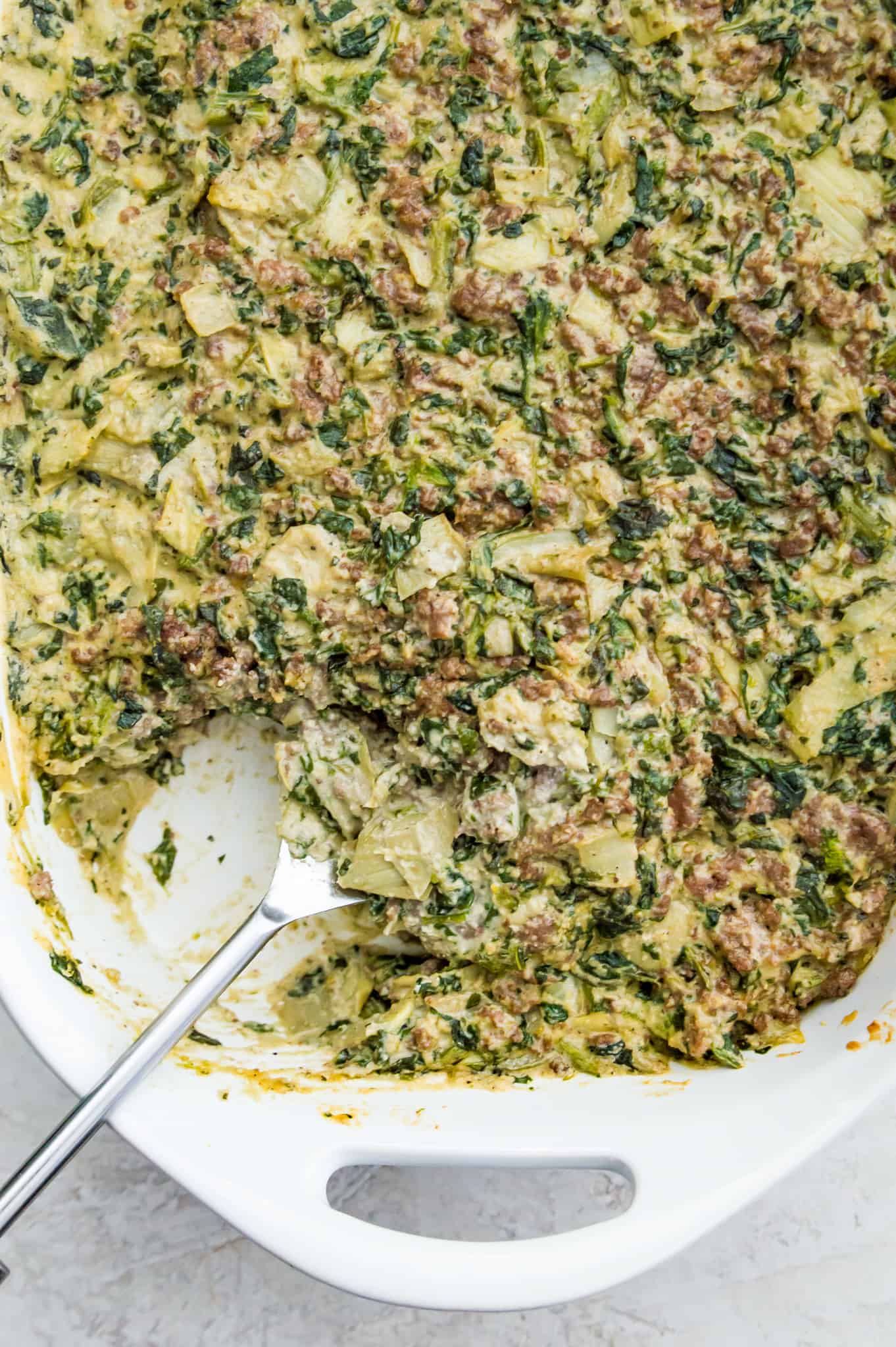 A pan of paleo spinach and artichoke casserole with a serving spoon in it.