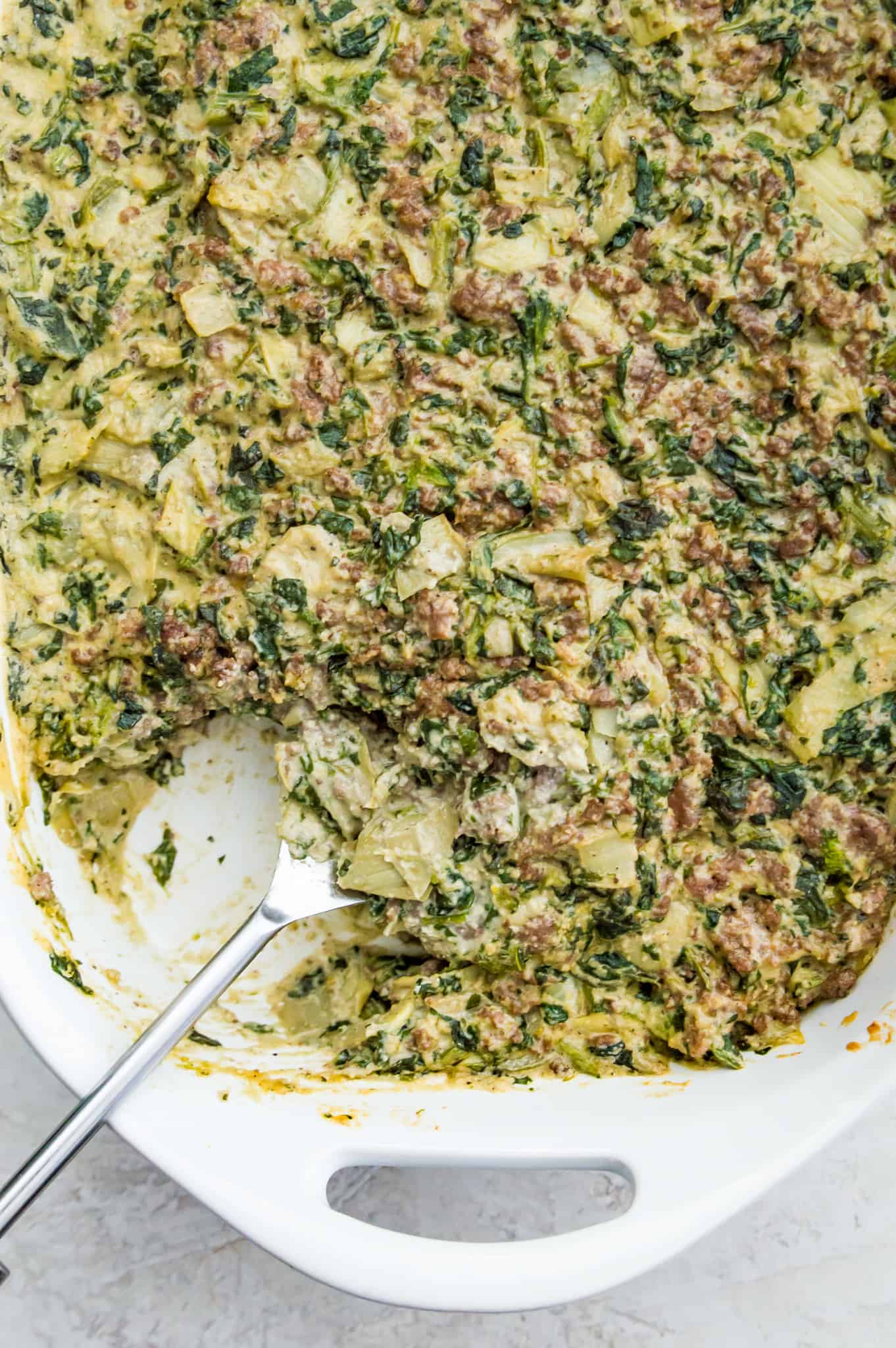 Paleo Spinach Beef Casserole A pan of paleo spinach and artichoke casserole with a serving spoon in it.