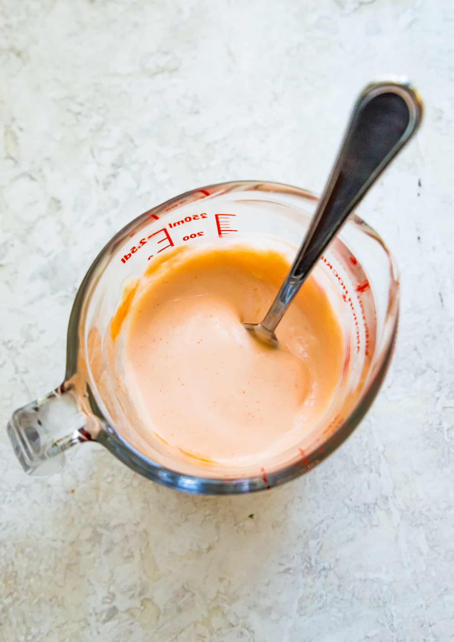 A glass measuring cup filled with sriracha mayo with a spoon in it.