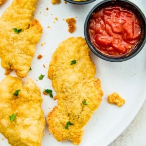 Cooked chicken tenders on a plate with a dish full of ketchup beside them.