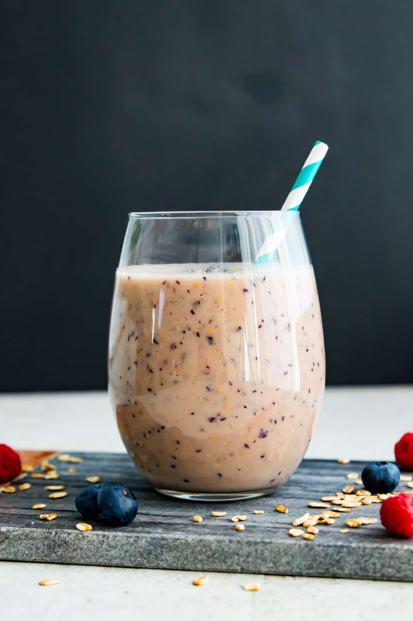 A clear glass full of a smoothie with a straw in it and surrounded by blueberries and raspberries. 