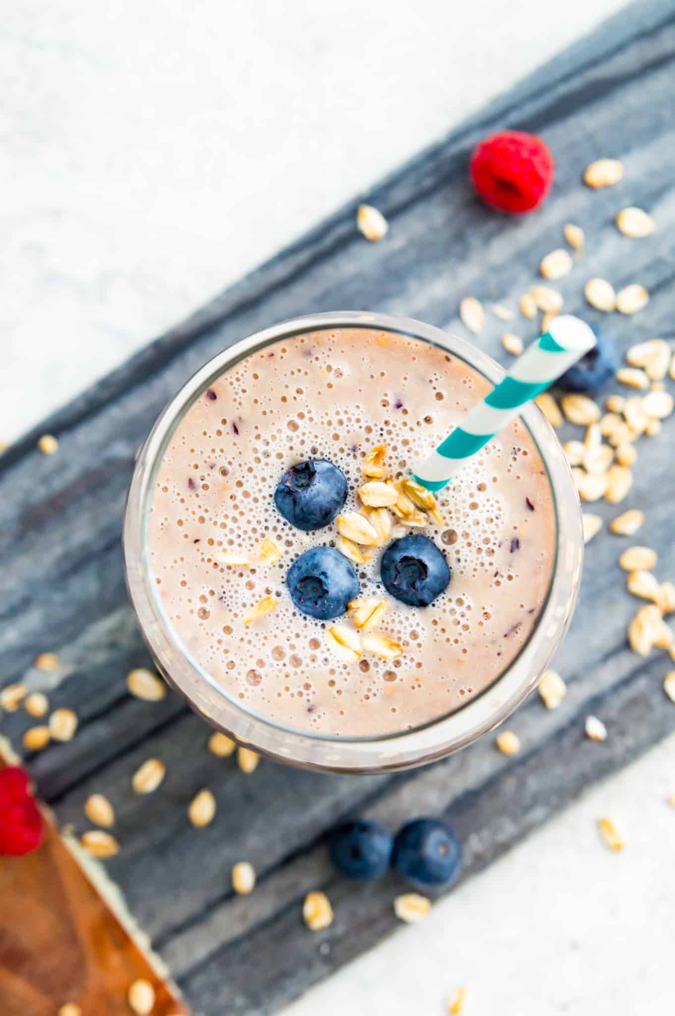 A glass of oat milk smoothie with a straw in it and blueberries on top.