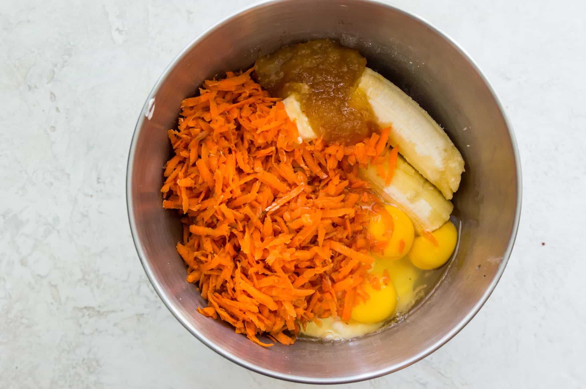 A mixing bowl with shredded carrots, eggs, bananas and sugar in it.