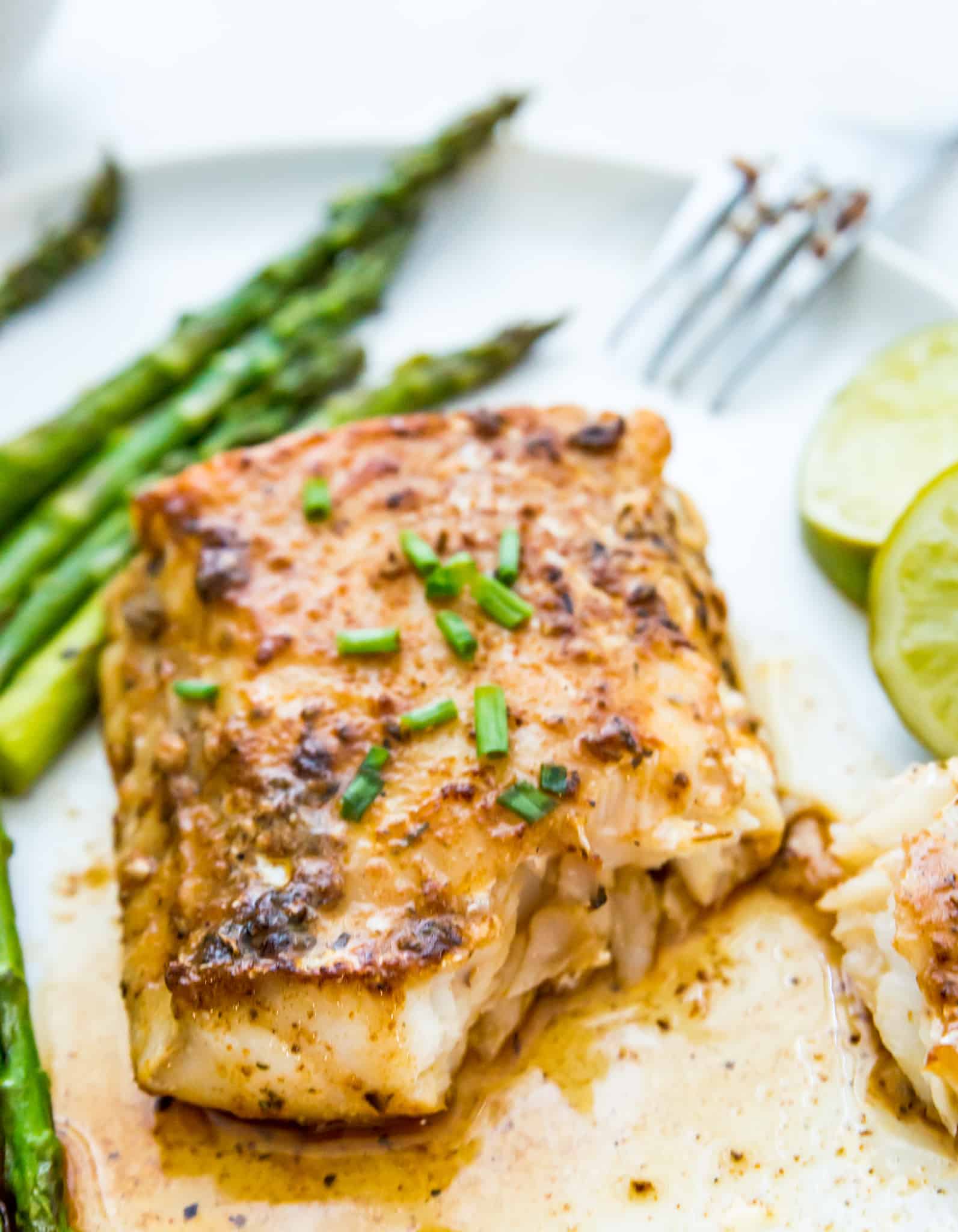 A piece of cooked cod on a plate with asparagus and a lime wedge next to it.