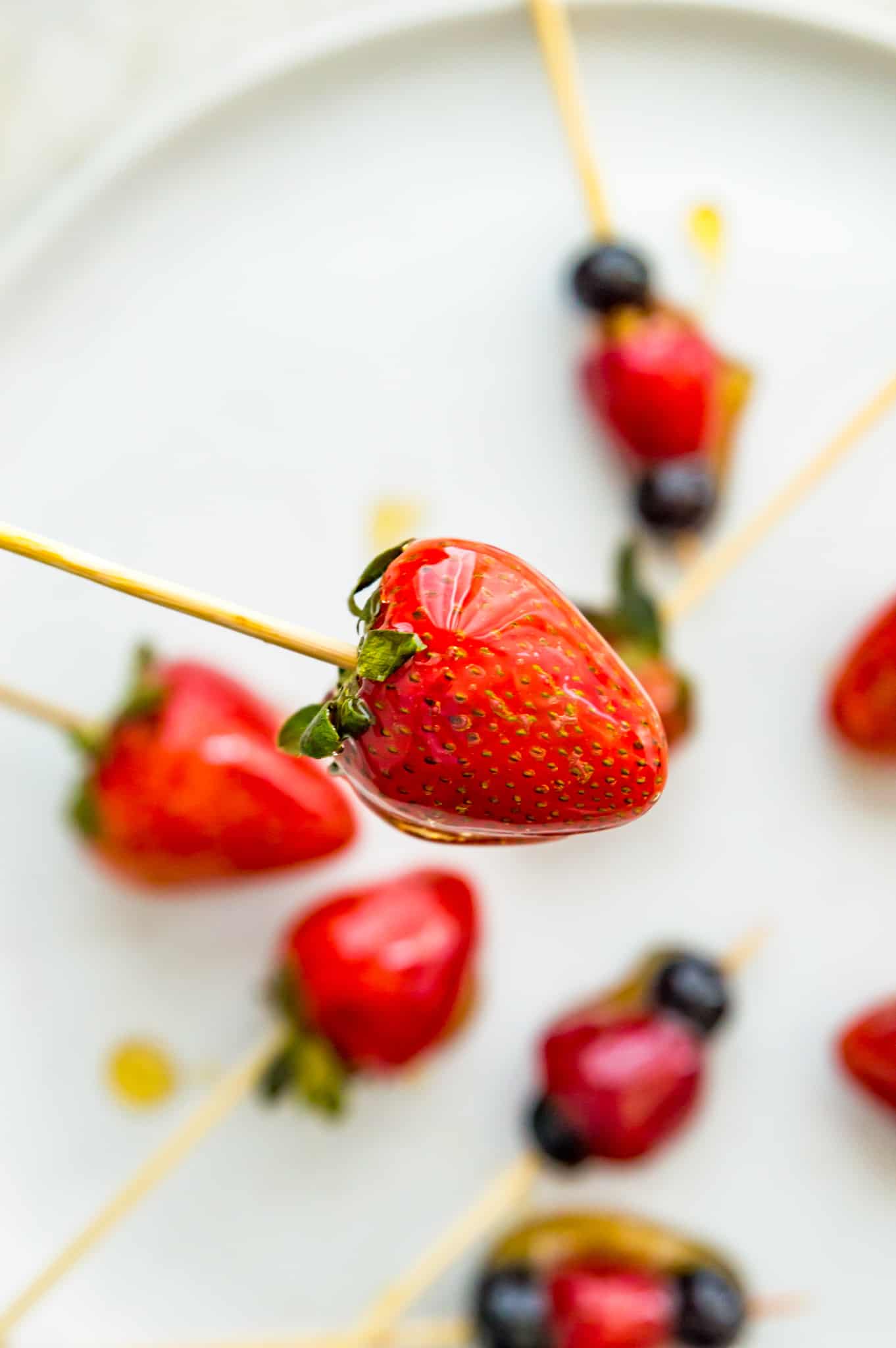 A Tanghulu strawberry on a bamboo skewer with a plate filled with candied strawberries below it.