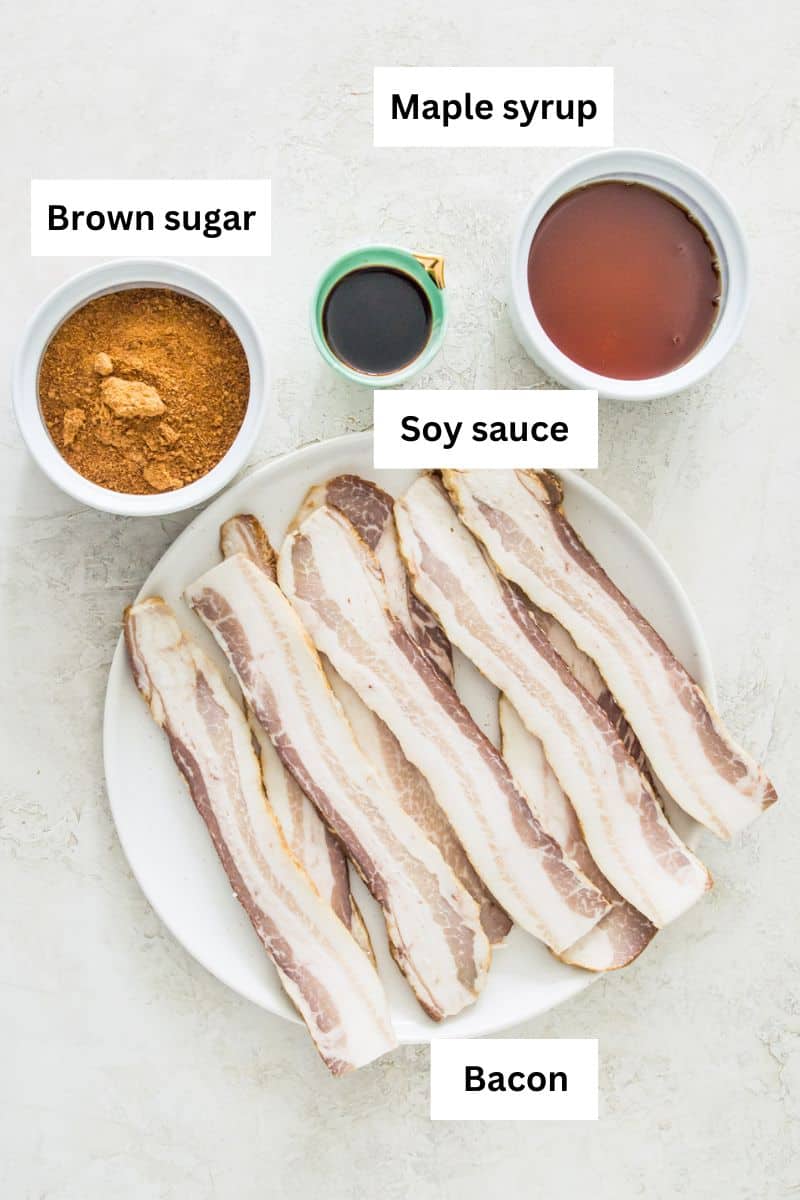 A plate with uncooked bacon on it, a bowl with brown sugar in it, a bowl with maple syrup in it and a smaller bowl with soy sauce in it.