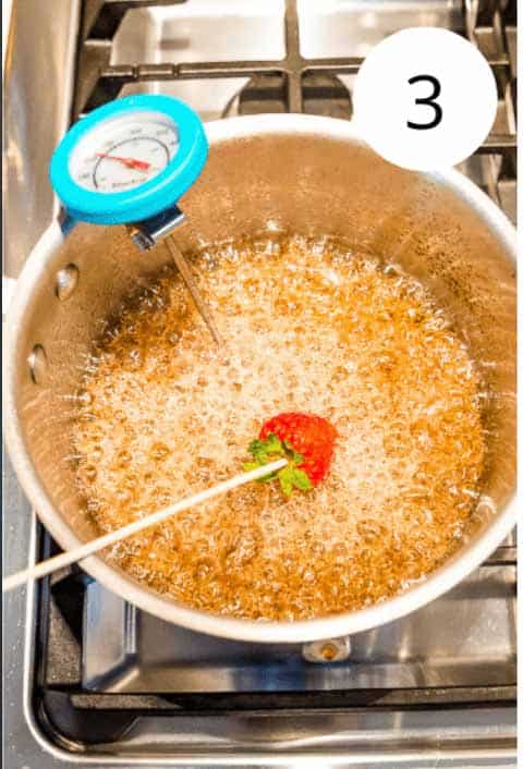 A strawberry on a bamboo skewer being dipping into a pot with boiling sugar water in it.