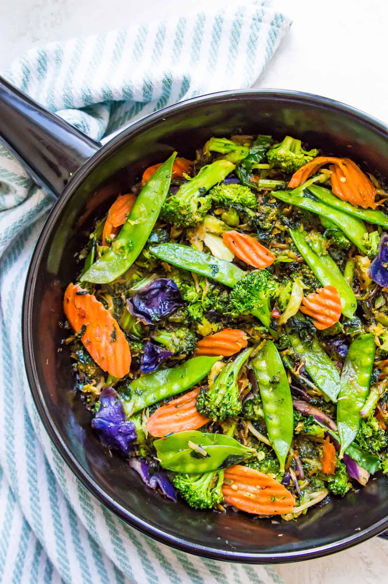 Stir fried asian vegetables in a black pan with a striped towel beside it.