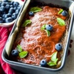 A loaf of meatloaf in a pan topped with barbecue sauce and garnished with blueberries and parsley leaves.