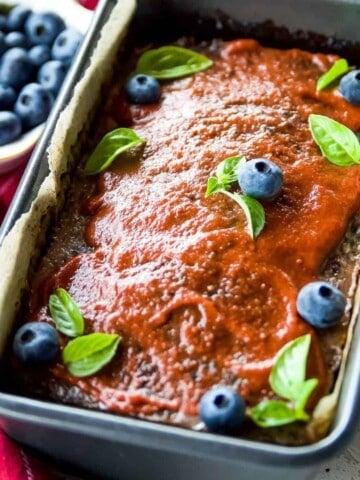A loaf of meatloaf in a pan topped with barbecue sauce and garnished with blueberries and parsley leaves.