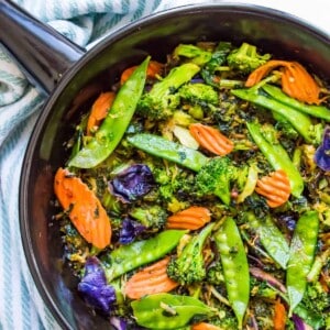 A black frying pan filled with stir fried asian vegetables including pea, carrots, broccoli and cabbage.