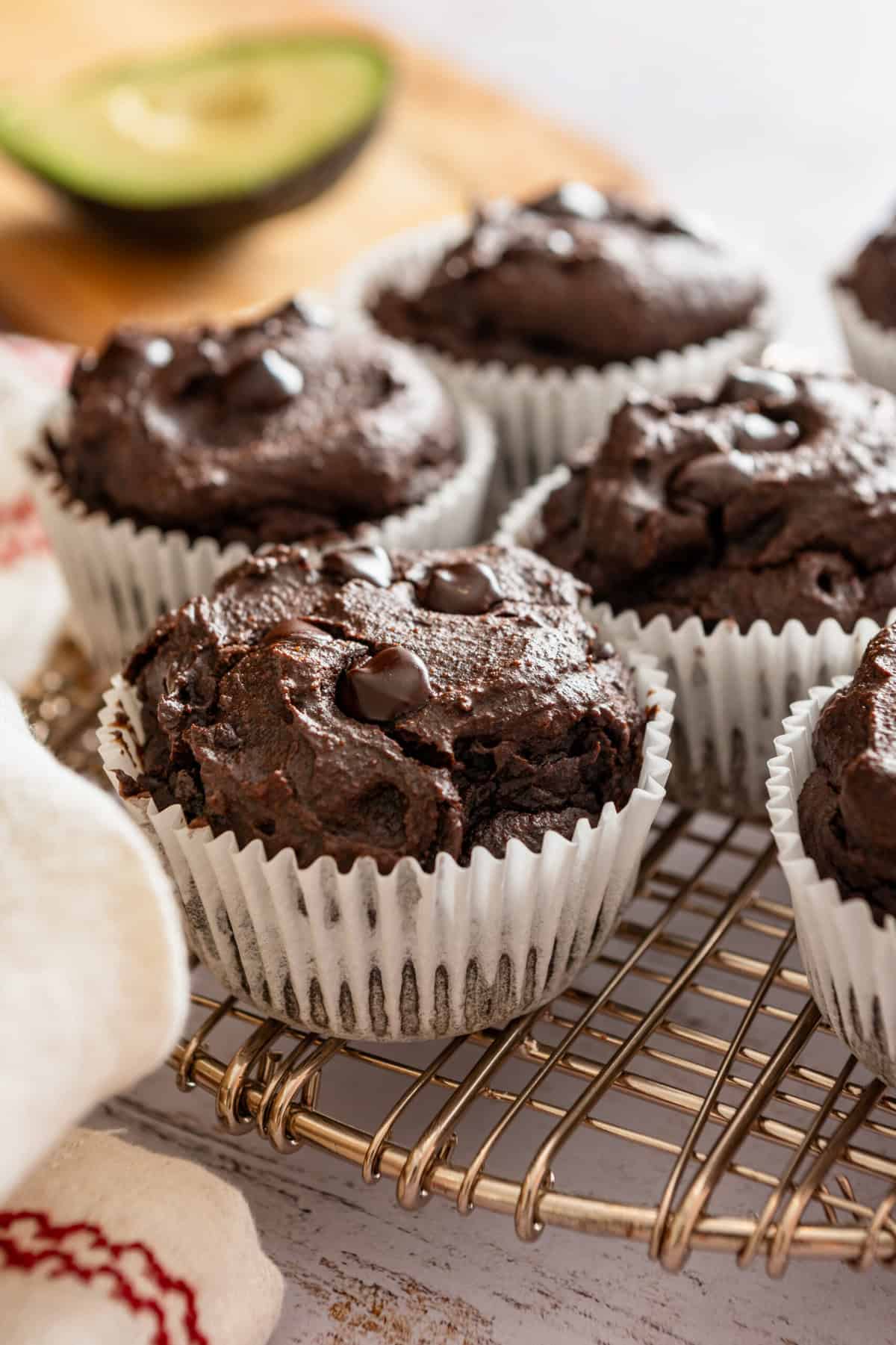 A batch of chocolate avocado muffins on a wire rack with an avocado next to them.
