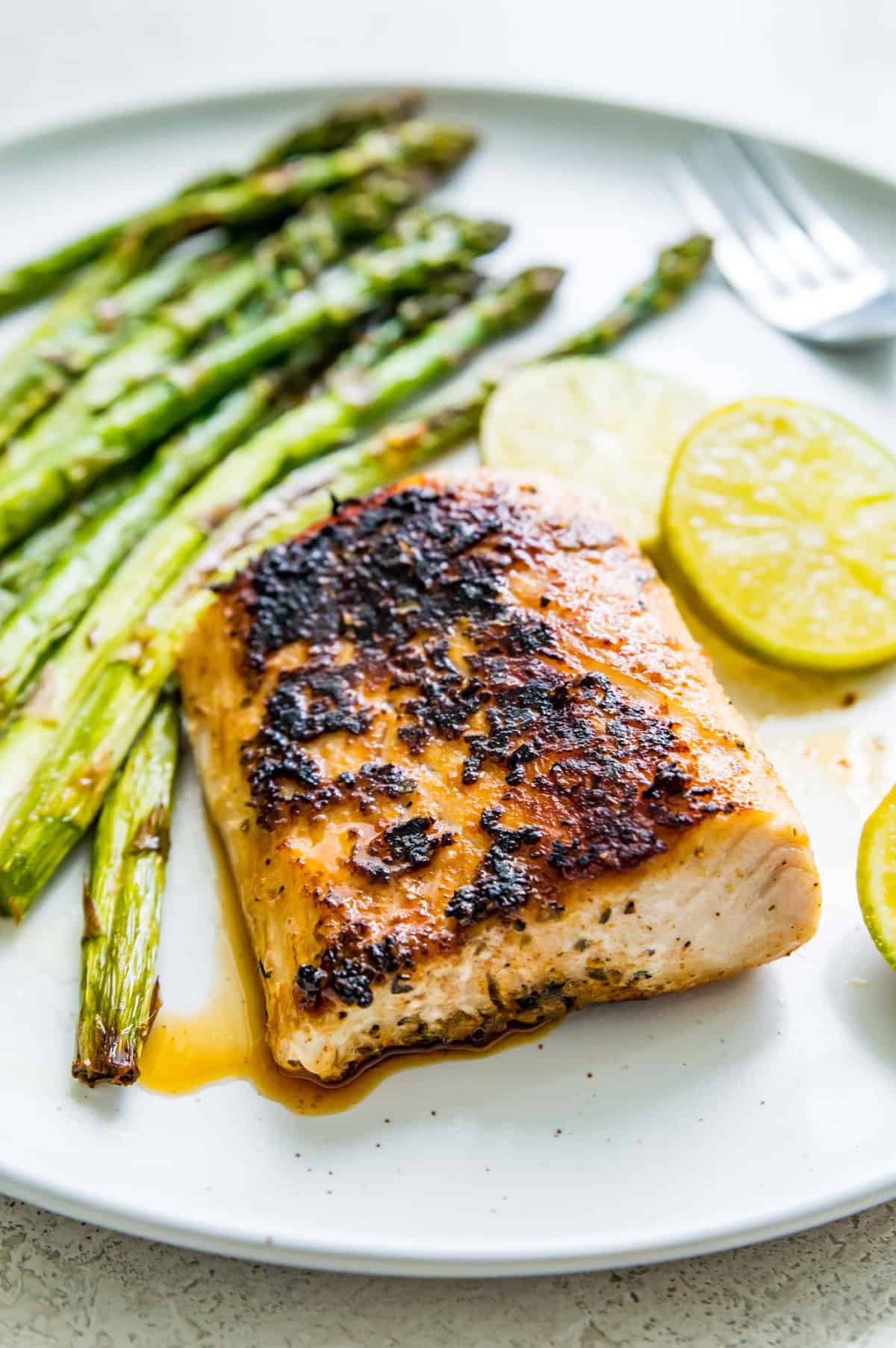 A piece of blackened Mahi Mahi on a plate with cooked asparagus and lemon wedges beside it.