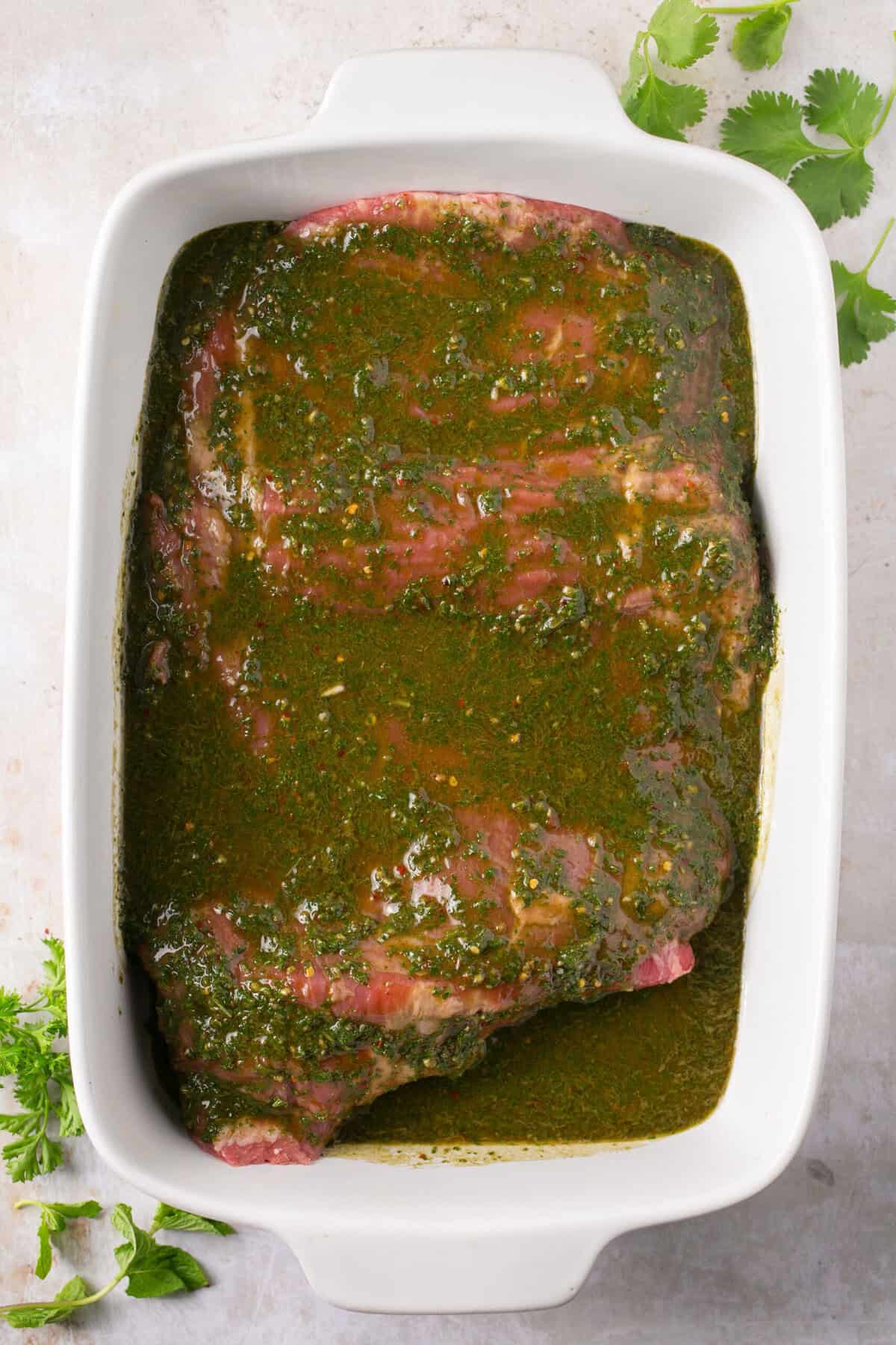 A piece of steak in a baking dish topped with chimichurri sauce.