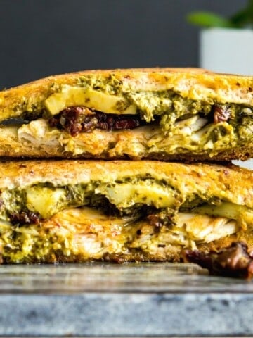 Two halves of a turkey pesto sandwich stacked on top of each other on a cutting board.