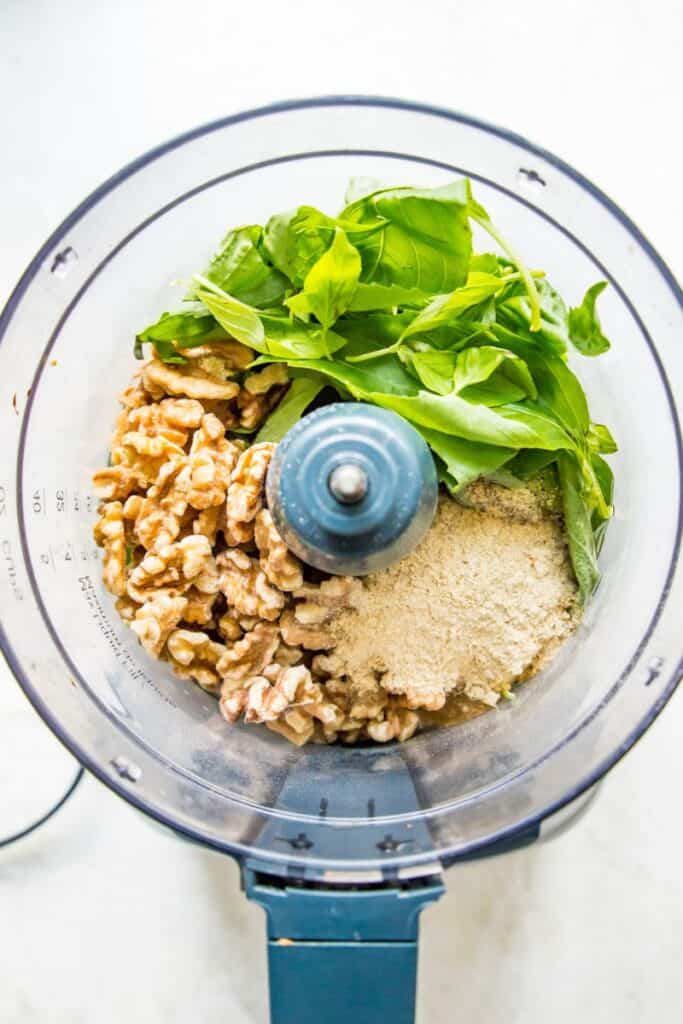 A food processor filled with fresh basil leaves, walnuts, avocado oil and nutritional yeast. 