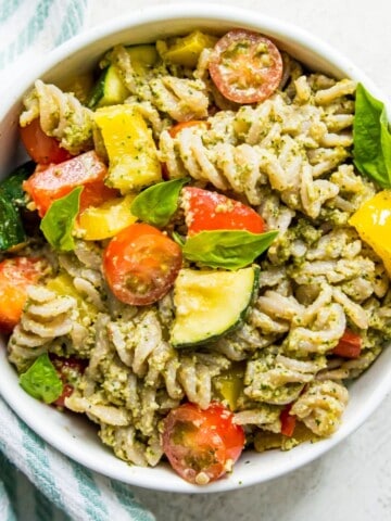 A bowl of veggie pesto pasta with chopped baby tomatoes and basil leaves on top.