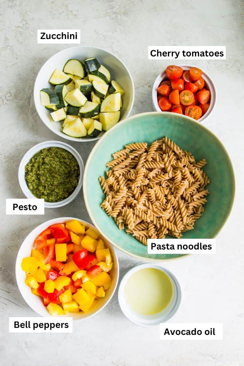 Bowls with different ingredients in them including pasta noodles, chopped zucchini, cherry tomatoes and pesto.