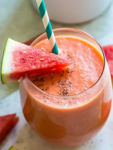 A pink smoothie in a glass with a piece of watermelon in it and a straw.