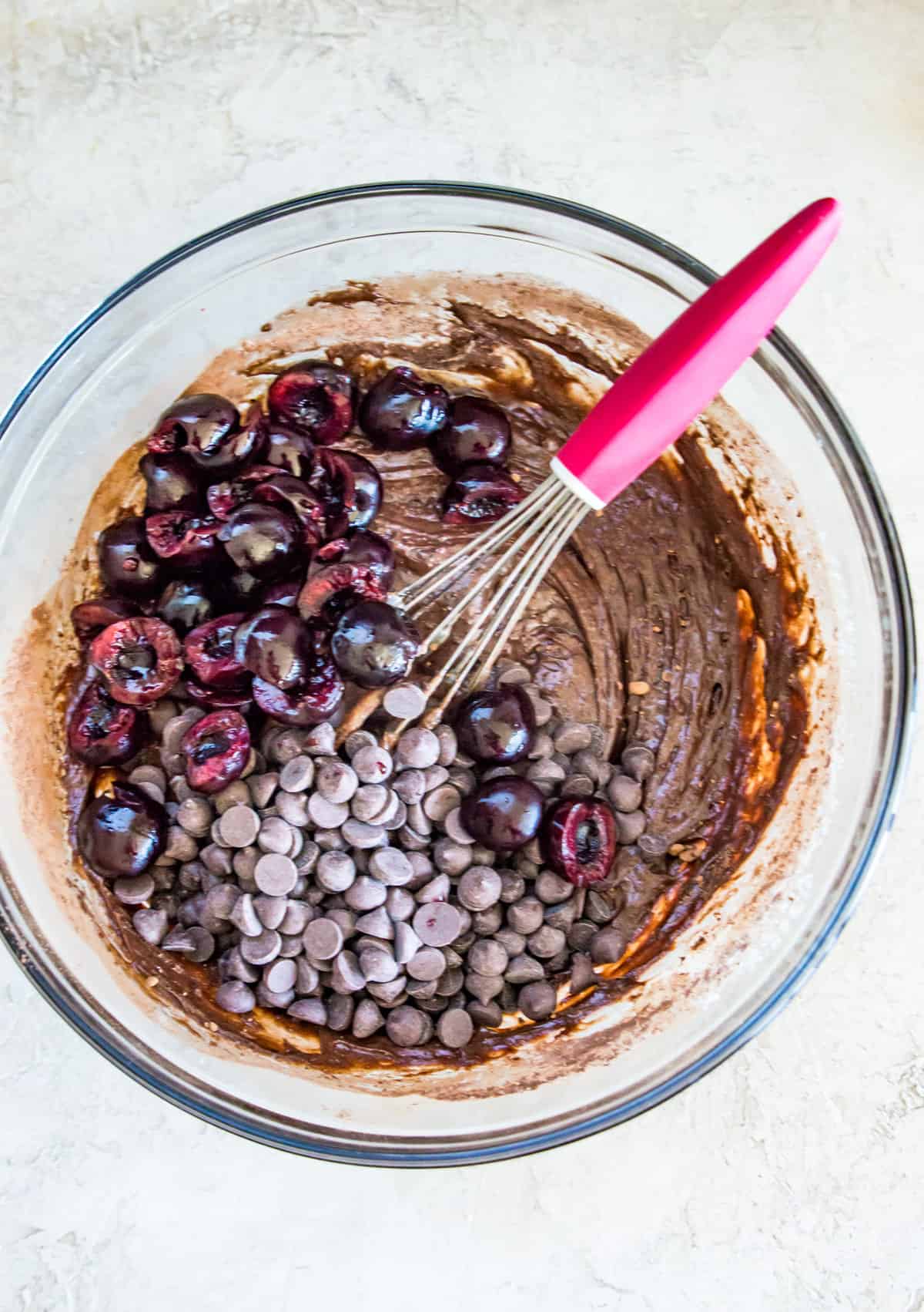 A glass bowl with pitted cherries, chocolate chips and a chocolate batter in it.