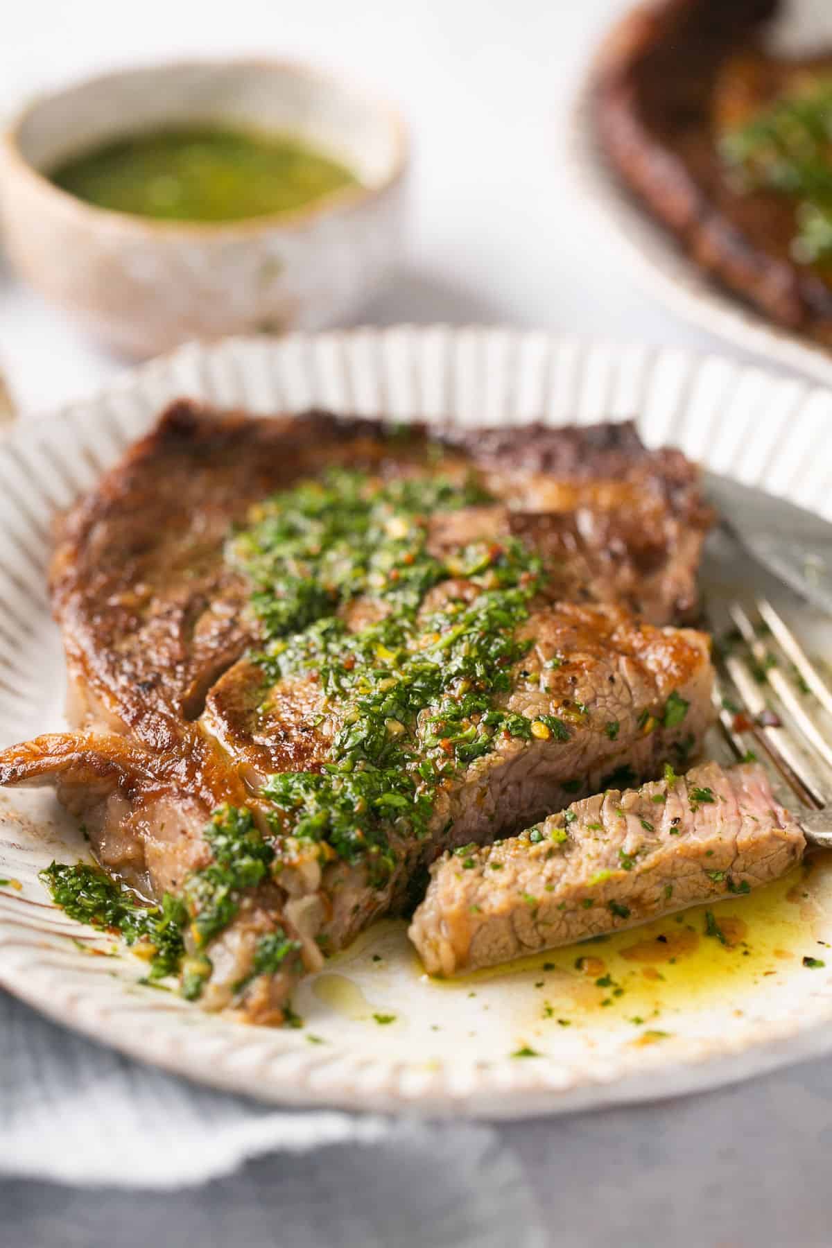 A piece of steak on a white plate with chimichurri sauce on it and a fork beside the steak.